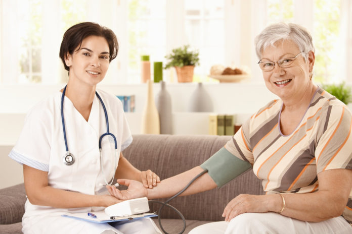 Nurse measuring blood pressure of senior woman at home. Looking at camera, smiling
